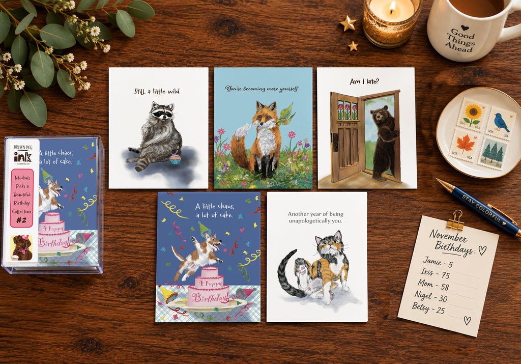 Styled flat lay of five illustrated animal birthday cards by Susan J. York with a matching card box, surrounded by flowers, stamps, a candle, mug, pen, and birthday reminder list.