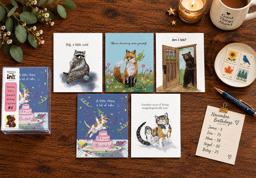 Styled flat lay of five illustrated animal birthday cards by Susan J. York with a matching card box, surrounded by flowers, stamps, a candle, mug, pen, and birthday reminder list.
