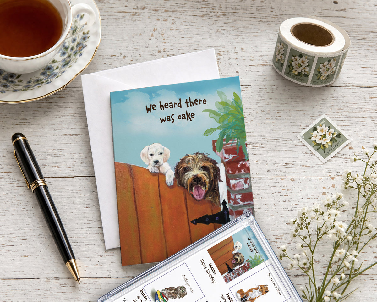 Brown Dog Ink birthday greeting card featuring two playful dogs peeking over a gate with “We heard there was cake,” shown with a white envelope, pen, tea, and postage stamps.