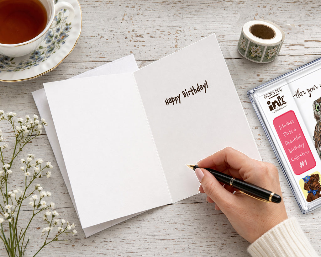 Open Brown Dog Ink birthday greeting card with “Happy Birthday!” inside and plenty of blank space for a personal handwritten message, shown with a pen, envelope, and Mocha’s Picks birthday card collection.