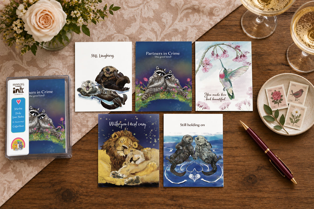 Love Notes & Anniversary greeting card collection featuring five illustrated animal cards with original artwork by Susan J. York, including otters, raccoons, hummingbird and lions, displayed with Brown Dog Ink packaging on a warm tabletop.