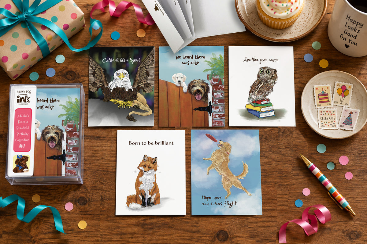 A styled flat lay of five illustrated animal birthday cards by Susan J. York with a matching card box on a wooden table, surrounded by colorful ribbons, confetti, a cupcake, stamps, a mug, and a striped pen.
