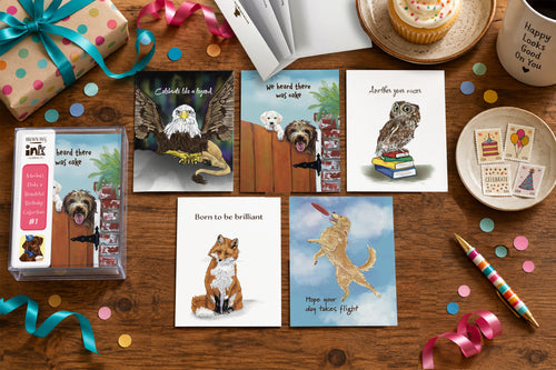 A styled flat lay of five illustrated animal birthday cards by Susan J. York with a matching card box on a wooden table, surrounded by colorful ribbons, confetti, a cupcake, stamps, a mug, and a striped pen.
